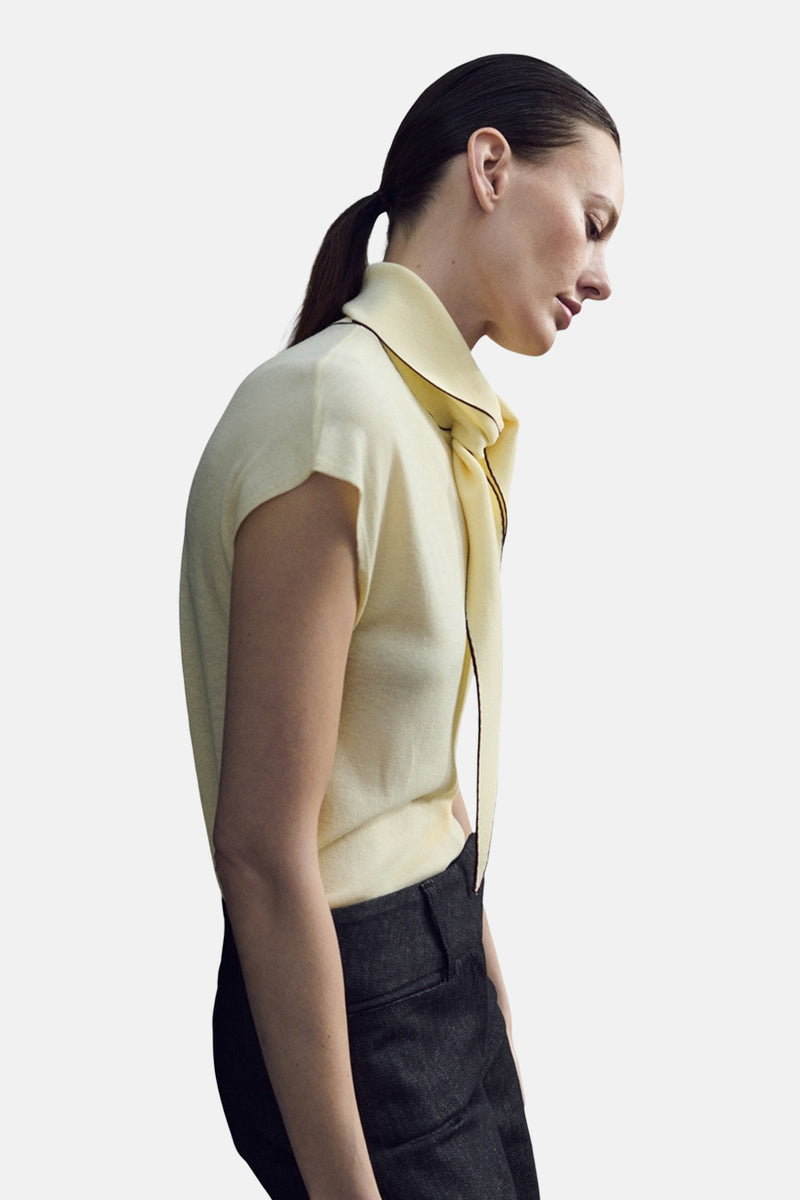 Chemisier jaune sans manches de MANGO, avec col montant et finitions contrastantes marron, porté avec un pantalon foncé.
