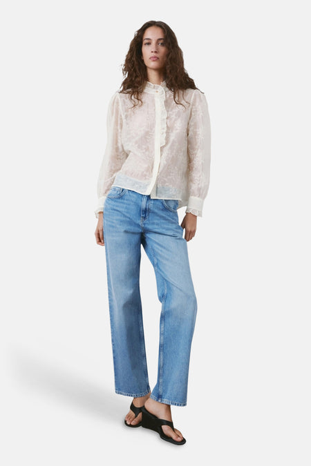 Chemisier beige à manches longues de MANGO, avec détails floraux délicats et col à volants, porté avec un jean bleu ample.

