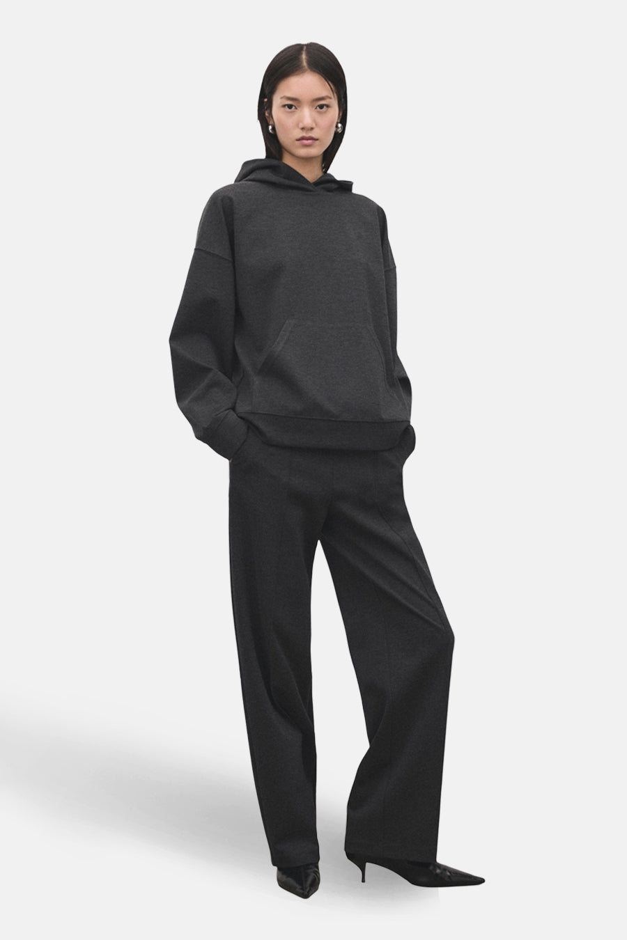 Pantalon gris de MANGO, avec coupe ample et taille haute, porté avec un sweat à capuche assorti et des escarpins noirs.