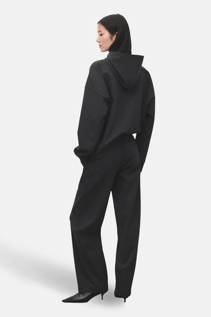 Pantalon gris de MANGO, coupe droite, porté avec un sweat à capuche et des escarpins noirs.