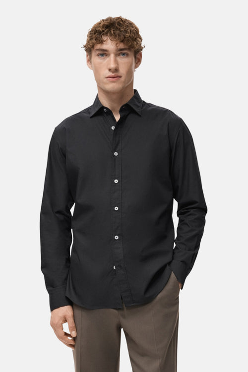 Chemise noire à manches longues de MANGO, portée avec un pantalon marron clair.