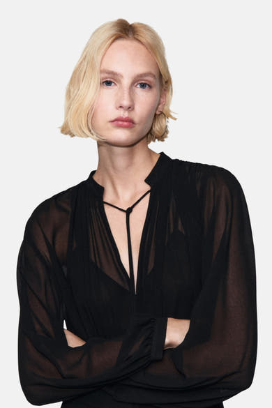 Zwarte jumpsuit van MANGO met diepe halslijn en doorschijnende lange mouwen, gedragen door een blond model.