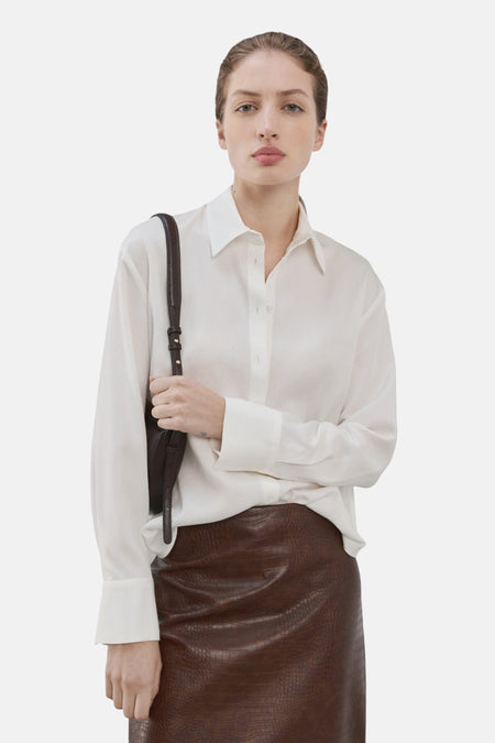 Chemise blanche à manches longues de MANGO, avec une fermeture boutonnée et un style décontracté. Portée avec une jupe en cuir marron.