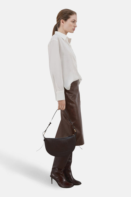 Chemise à manches longues blanche de MANGO, portée avec une jupe marron texturée et des bottes assorties.
