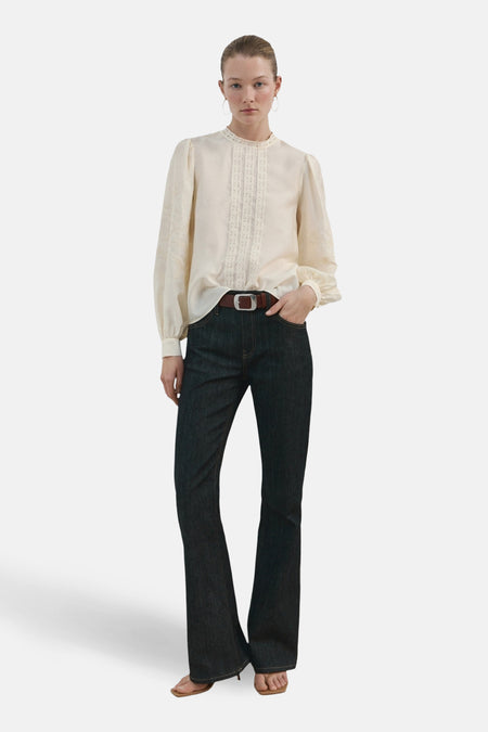 Chemisier écru (manches longues) de MANGO, orné de dentelle verticale et porté avec un jean taille haute et une ceinture marron.
