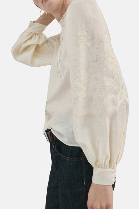 Chemisier écru à manches longues de MANGO, avec broderies florales et porté avec un jean foncé et une ceinture marron.