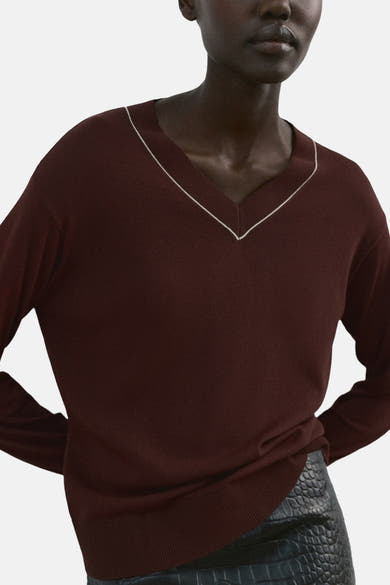 Pull à col en V brun de MANGO, orné d'une bordure blanche contrastante au col, porté avec une jupe texturée.
