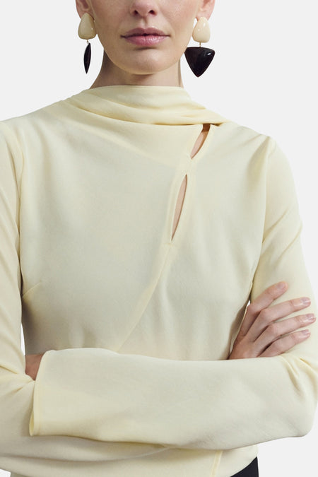 Blouse jaune à manches longues de MANGO, avec un col montant asymétrique et des boucles d'oreilles géométriques.
