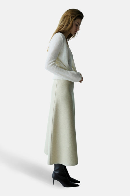 Gilet écru de MANGO, porté avec une jupe longue beige et des bottines noires à talons aiguilles.
