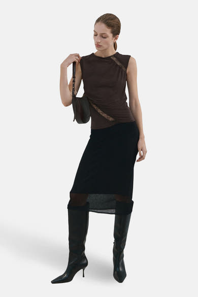 Chemisier sans manches brun MANGO avec détails en dentelle, porté avec une jupe noire transparente et des bottes en cuir.