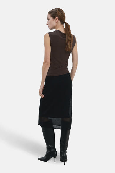 Blouse sans manches brun de MANGO, portée avec une jupe noire semi-transparente et des bottes en cuir noir à talons aiguilles.