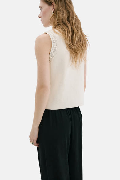 Gilet beige de MANGO, tricoté avec détails ajourés aux emmanchures, porté avec un pantalon noir ample.