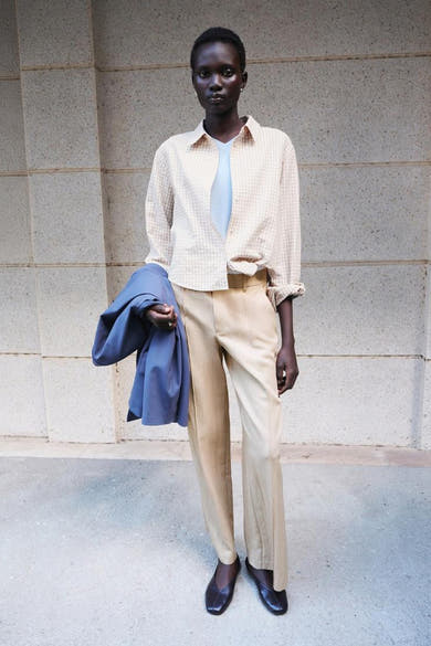 Beige geruite blouse van MANGO, open over een lichtblauwe top, opgerolde mouwen, gedragen met een beige broek en een blauwe jas in de hand.