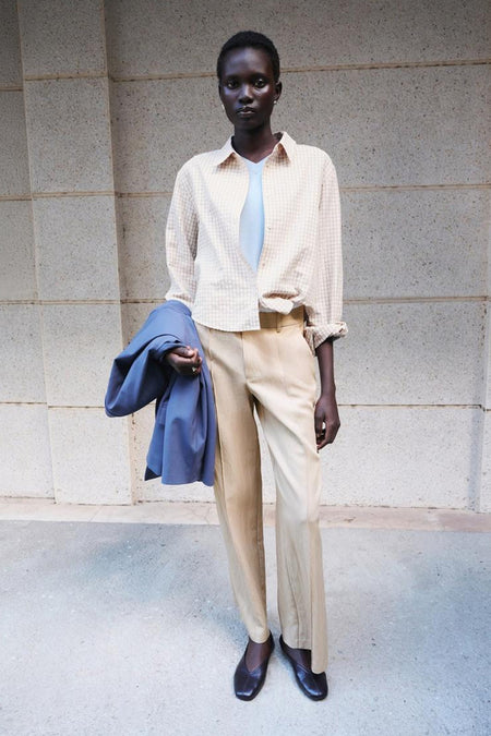 Chemise beige à carreaux de MANGO, ouverte sur un top bleu clair, manches retroussées, portée avec un pantalon beige et une veste bleue à la main.