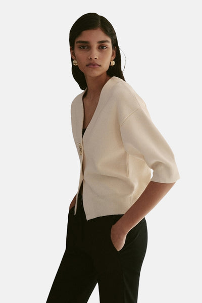 Gilet beige de MANGO, porté avec un pantalon noir et des boucles d'oreilles dorées.
