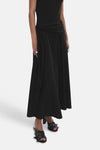 Jupe longue noire de MANGO, fluide, portée avec un haut noir noué à la taille, des sandales à boucles et des bagues.