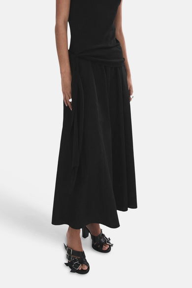 Jupe longue noire de MANGO, fluide, portée avec un haut noir noué à la taille, des sandales à boucles et des bagues.