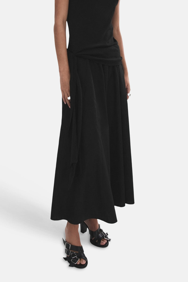 Jupe longue noire de MANGO, fluide, portée avec un haut noir noué à la taille, des sandales à boucles et des bagues.