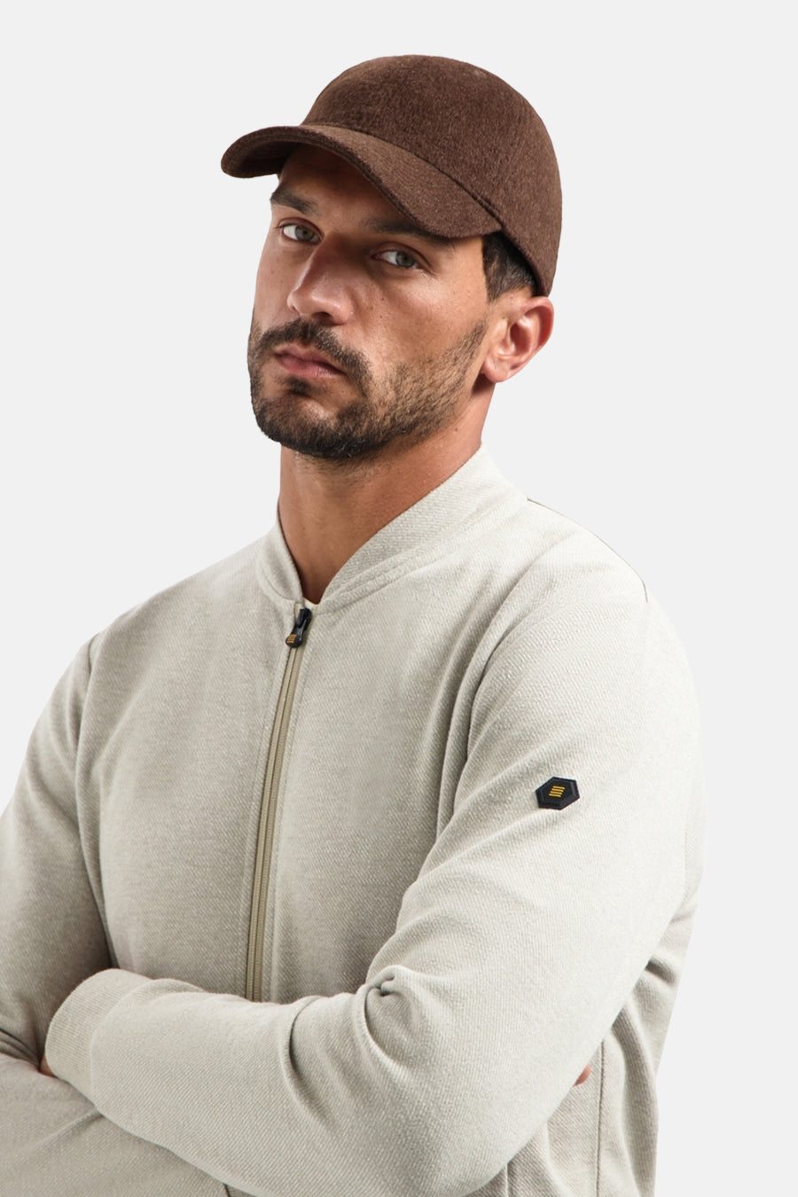 Beige gilet van NO EXCESS, met ritssluiting en zwart embleem op de mouw, gedragen met een bruine pet.