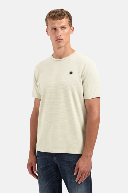 Beige gestreept T-shirt met korte mouwen van NO EXCESS, met een zwart embleem op de borst, gedragen met een blauwe spijkerbroek.