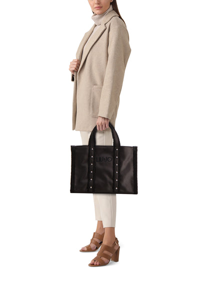 Sac à bandoulière noir de LIU JO, avec garniture en fourrure et détails cloutés, porté par une femme en manteau beige.
