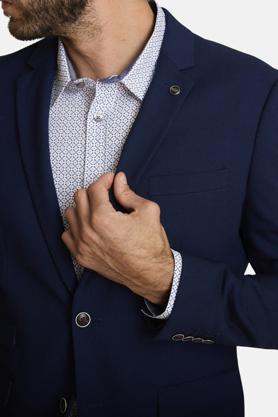 Blauwe blazer van DELAHAYE met een subtiele textuur, gedragen over een overhemd met geometrisch patroon en zichtbare knopen aan de mouwen.