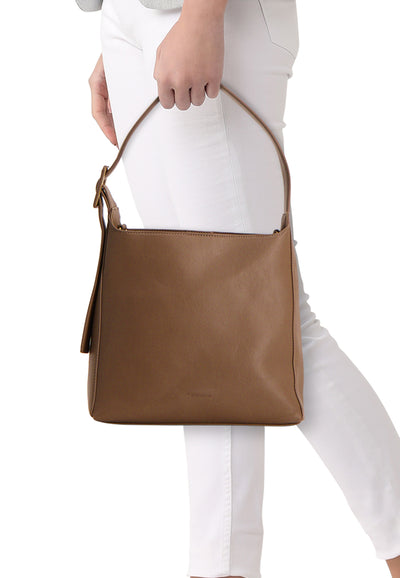 Sac à bandoulière taupe de TAMARIS, porté à l'épaule avec un pantalon blanc.