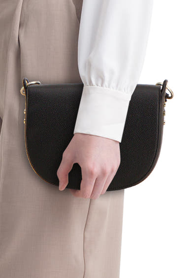 Zwarte handtas van TAMARIS, in de hand gehouden met een wit overhemd en een beige broek.