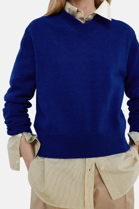 Pull col V bleu de MANGO, tricot fin, porté sur chemise à fines rayures jaunes, vue de face.