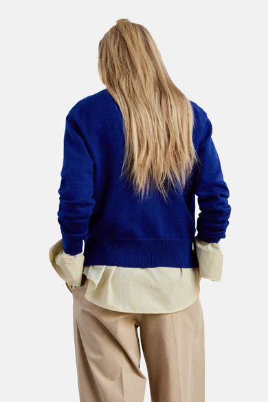 Pull col V bleu de MANGO, vue dos, tricot uni, manches longues, porté sur chemise à fines rayures et pantalon beige