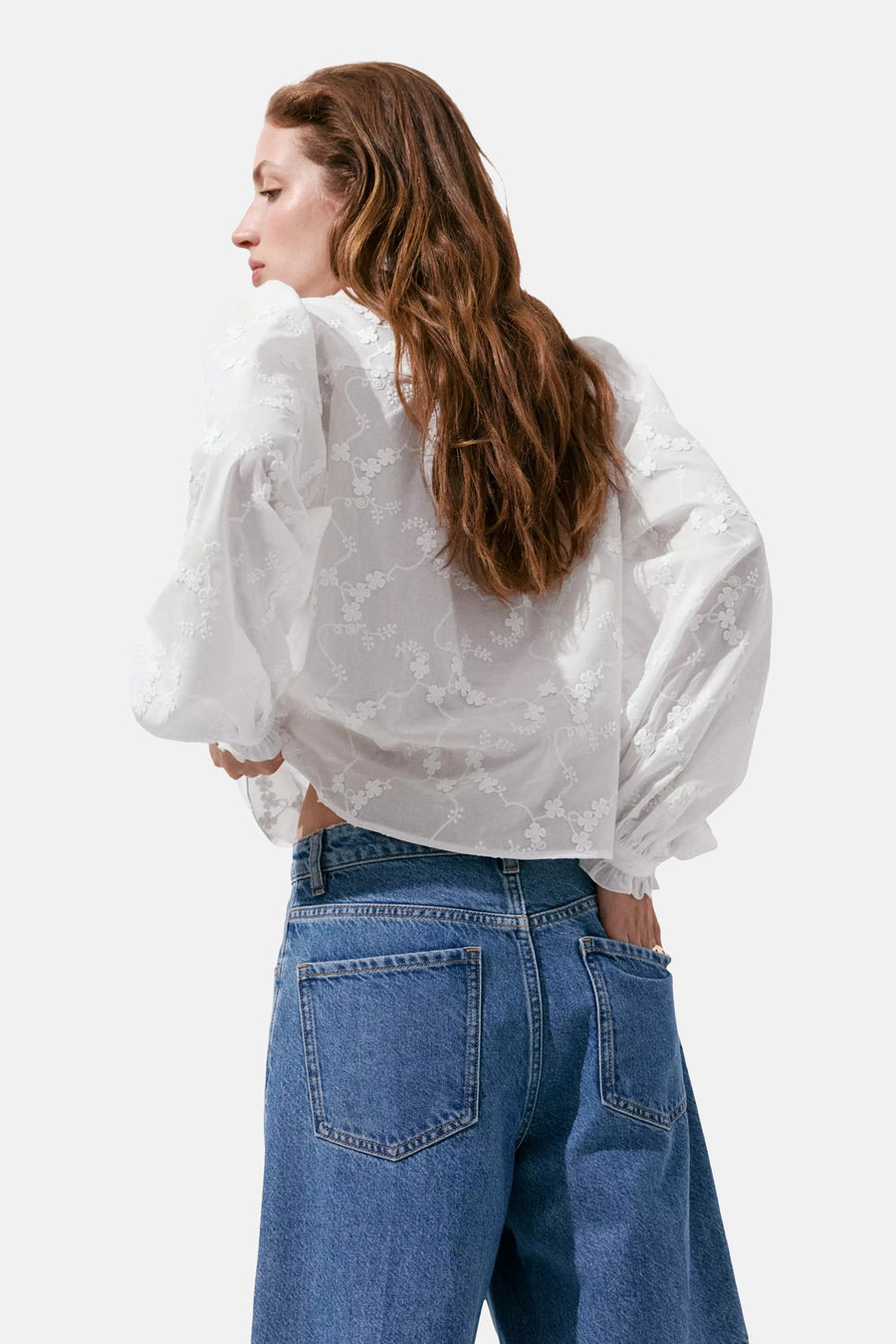 White long-sleeve blouse with floral embroidery and ruffled sleeves, shown from back view on model wearing blue jeans.