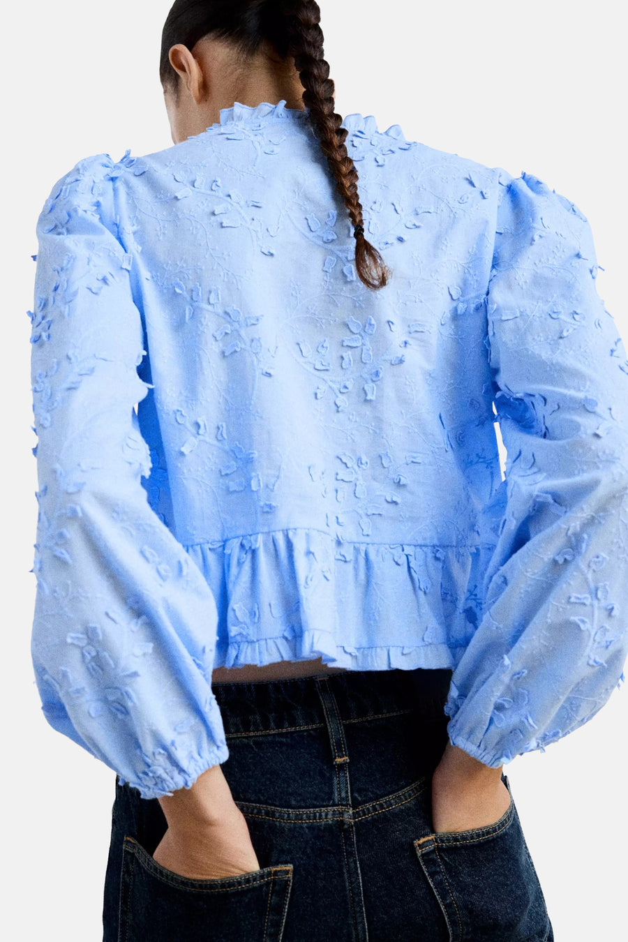 Light blue long-sleeve blouse with floral texture and ruffled hem, back view, worn with dark denim jeans