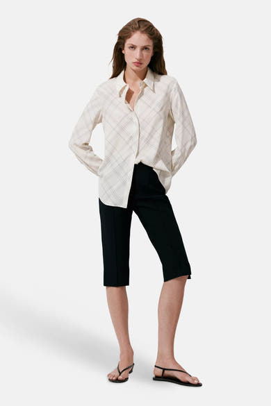 Cream long-sleeve shirt with thin black plaid pattern, worn open over black cropped pants and black sandals, full-length front view.