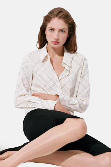Cream long-sleeve blouse with black grid pattern, worn by model in seated pose with black pants.