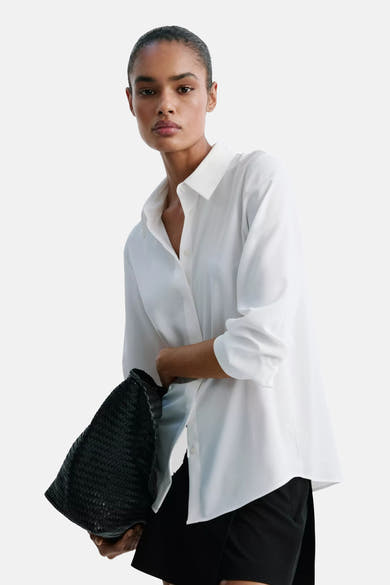 White long-sleeve blouse with classic collar and button front, worn open over black shorts, holding black woven bag, side view.