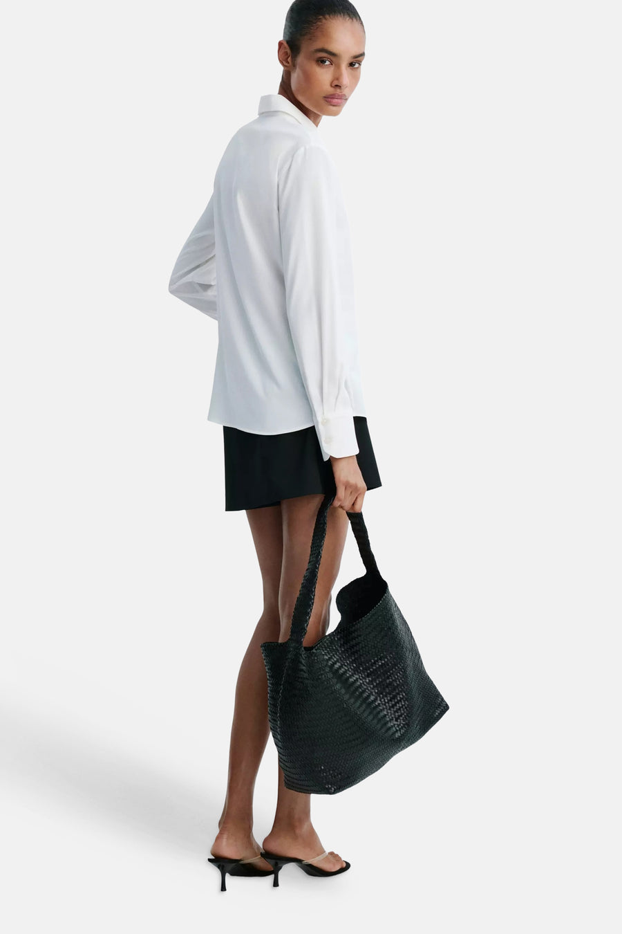 White long-sleeve blouse with classic collar and button cuffs, side view on model wearing black skirt and holding woven tote bag