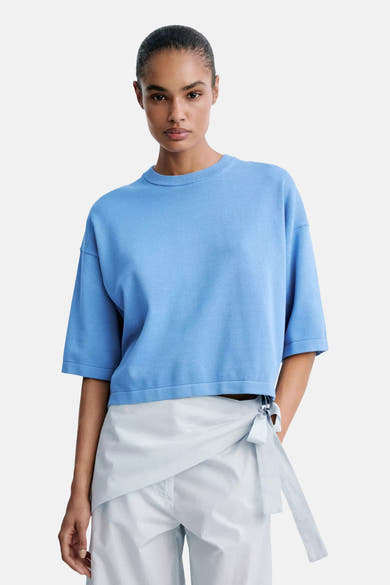 Light blue short-sleeve knit sweater with ribbed crew neck and cropped fit, worn with white wrap pants, front view