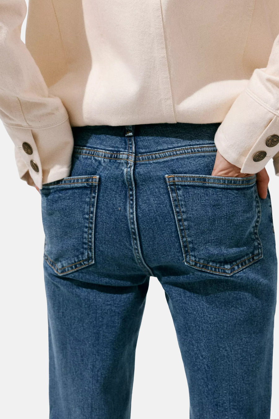 Mid blue denim flared jeans with classic back pockets and orange stitching, shown from rear view on model wearing cream top.