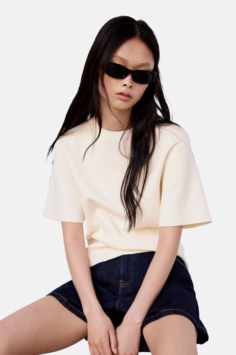 Ecru short-sleeve knit top with relaxed fit and round neckline, worn with dark denim shorts and black sunglasses, front view.