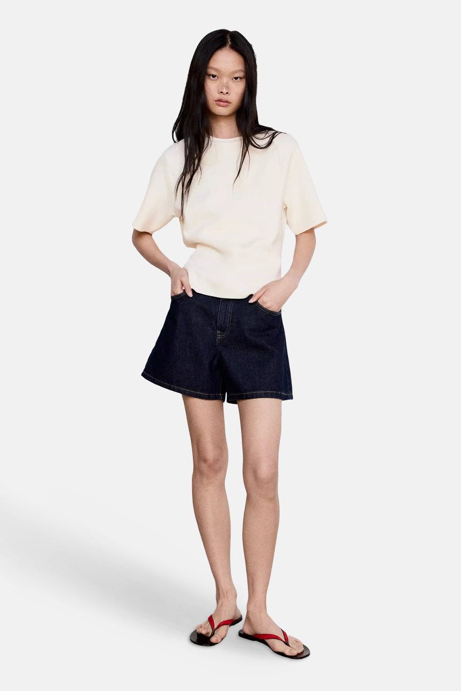 Ecru short-sleeve knit top with relaxed fit and round neckline, worn with dark denim shorts and red flip-flops, full-length front view