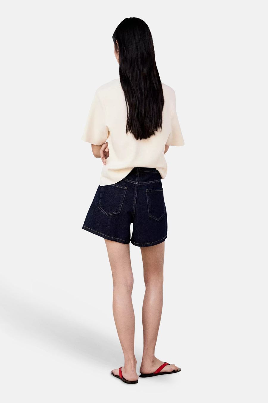 Ecru short-sleeve knit top from back view, soft texture, relaxed fit, worn with dark denim shorts and red sandals