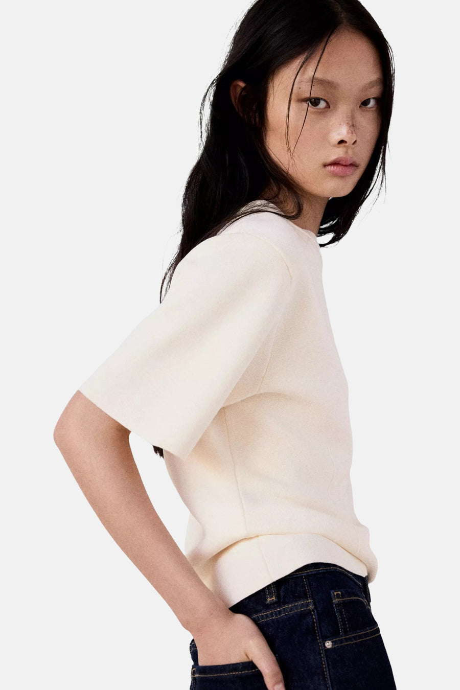 Ecru short-sleeve knit top with ribbed texture, side view on model wearing dark denim jeans