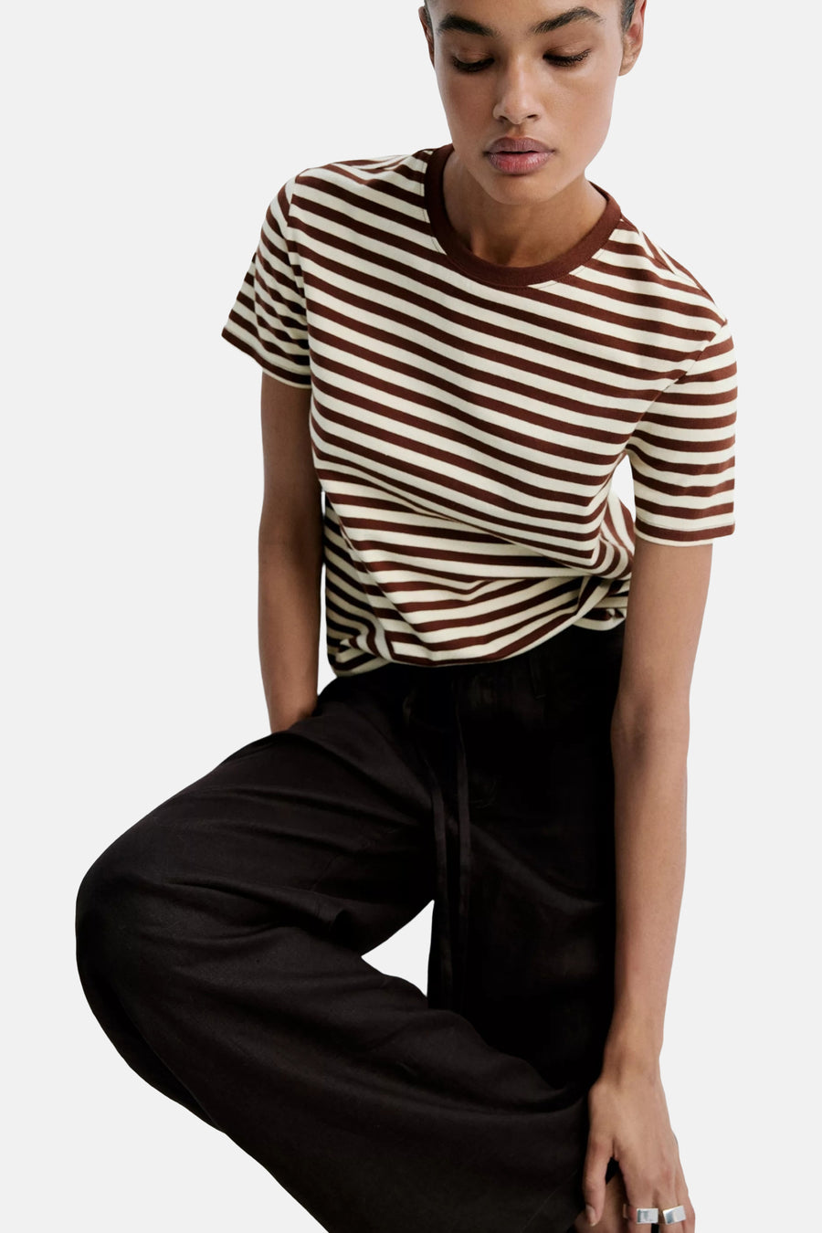 Multicolored short-sleeve striped t-shirt with brown and cream horizontal stripes, crew neck, worn by model in lifestyle pose with black pants.