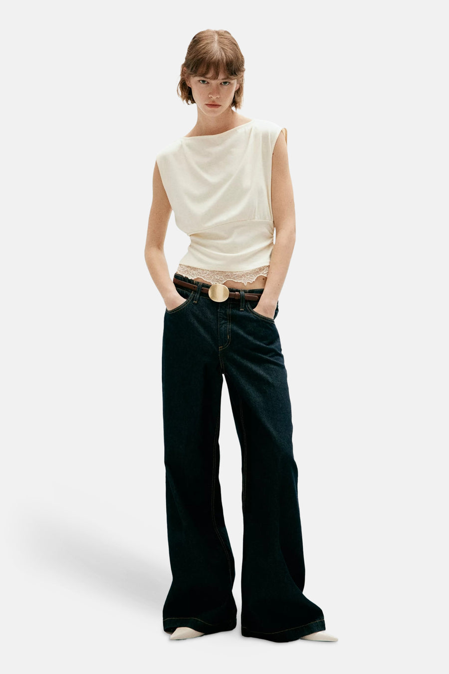Écru sleeveless top with asymmetrical neckline and draped front, worn with dark wide-leg jeans and brown belt, full-length lifestyle view