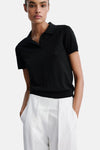 Black short-sleeve polo shirt with ribbed collar and cuffs, worn by model with white wide-leg pants, front view