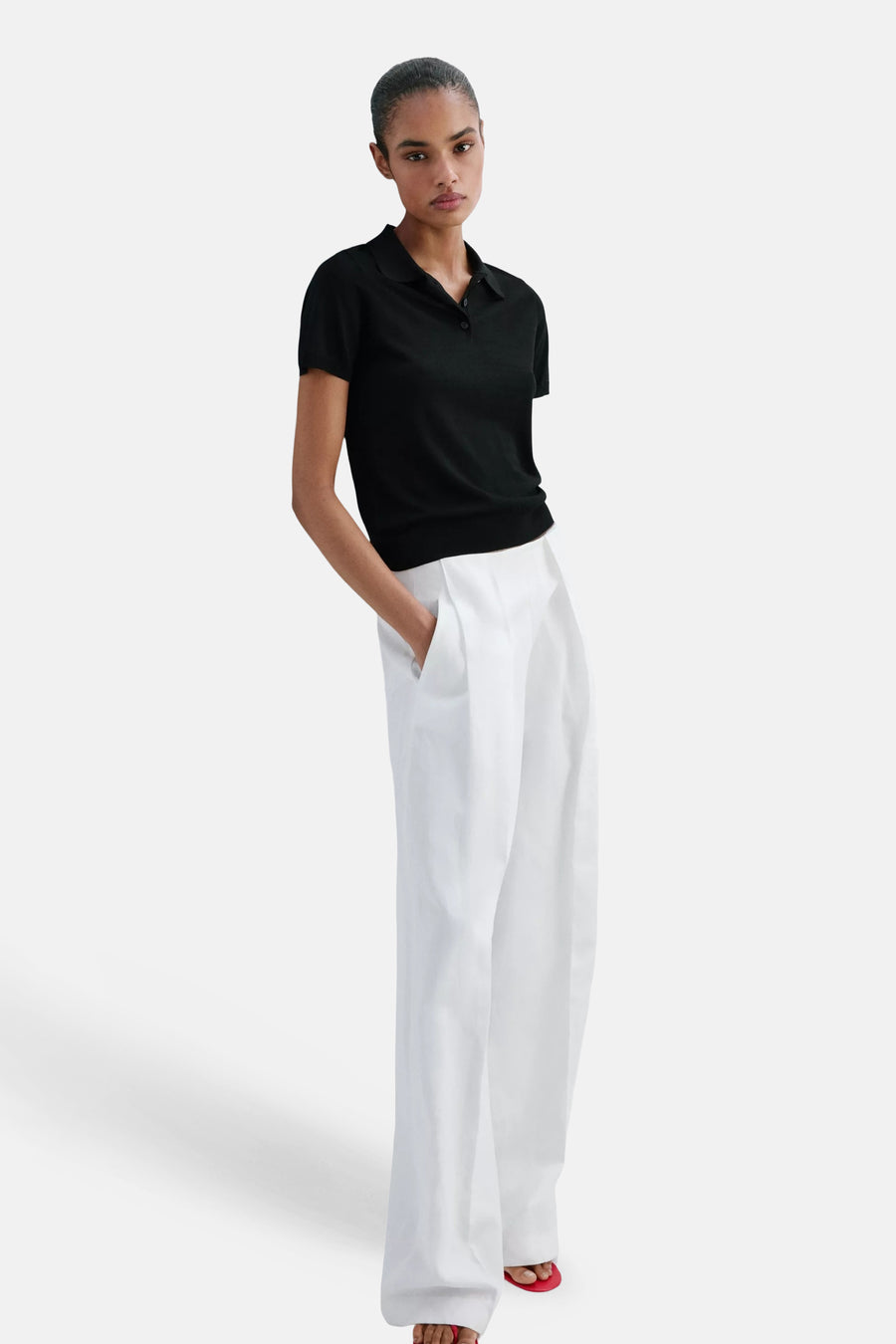 Black short-sleeve polo shirt with ribbed collar and button placket, worn with white wide-leg pants and red sandals, front view.