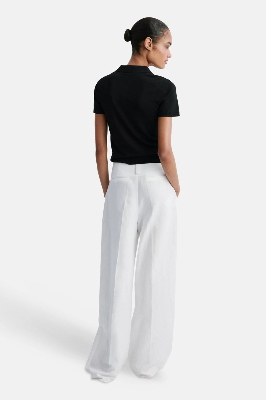 Black short-sleeve polo shirt from back view, fitted with classic collar, paired with white wide-leg trousers