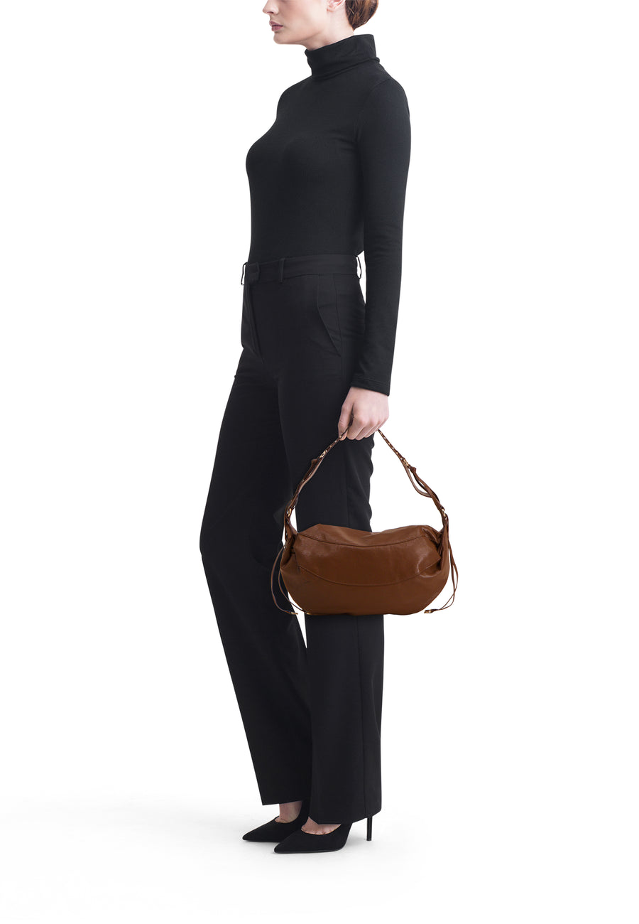 Brown leather shoulder bag with curved silhouette and braided strap, worn by model in black turtleneck and trousers, side view.