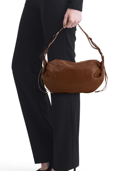 Brown leather shoulder bag with curved silhouette and braided strap, worn by model in black trousers, side view