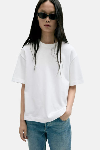 White short-sleeve t-shirt with relaxed fit and round neckline, worn by model in lifestyle front view with blue jeans and sunglasses.